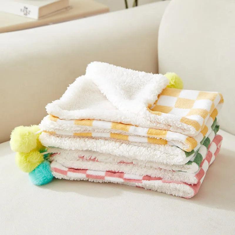 Green plaid pet blanket with fur ball trim on a sofa with a cat resting on it.
