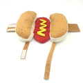 Small dog wearing a red and brown plush hot dog costume with bun and sausage design.