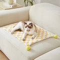 Green plaid pet blanket with fur ball trim on a sofa with a cat resting on it.