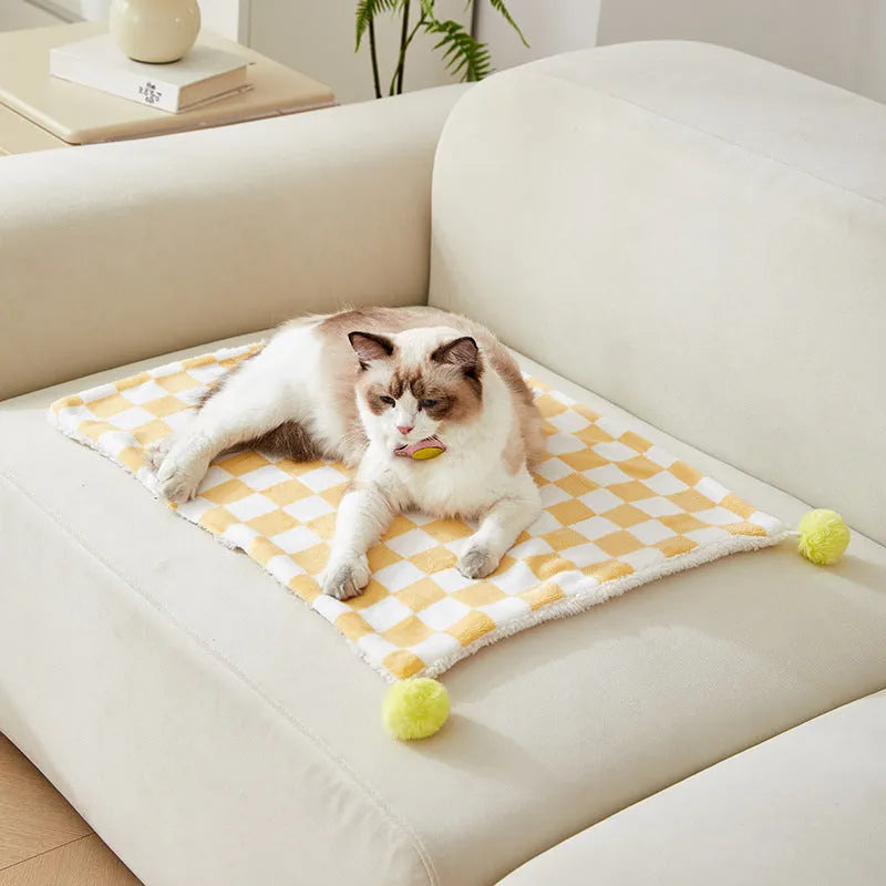 Green plaid pet blanket with fur ball trim on a sofa with a cat resting on it.