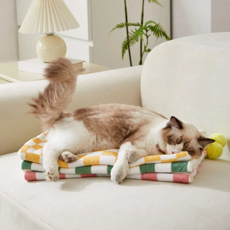 Green plaid pet blanket with fur ball trim on a sofa with a cat resting on it.