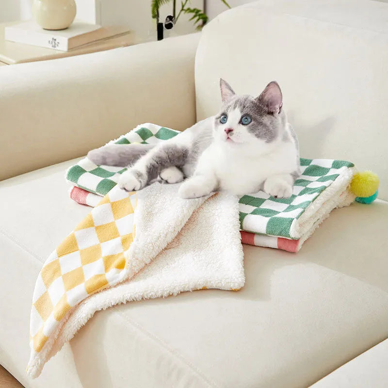 Green plaid pet blanket with fur ball trim on a sofa with a cat resting on it.