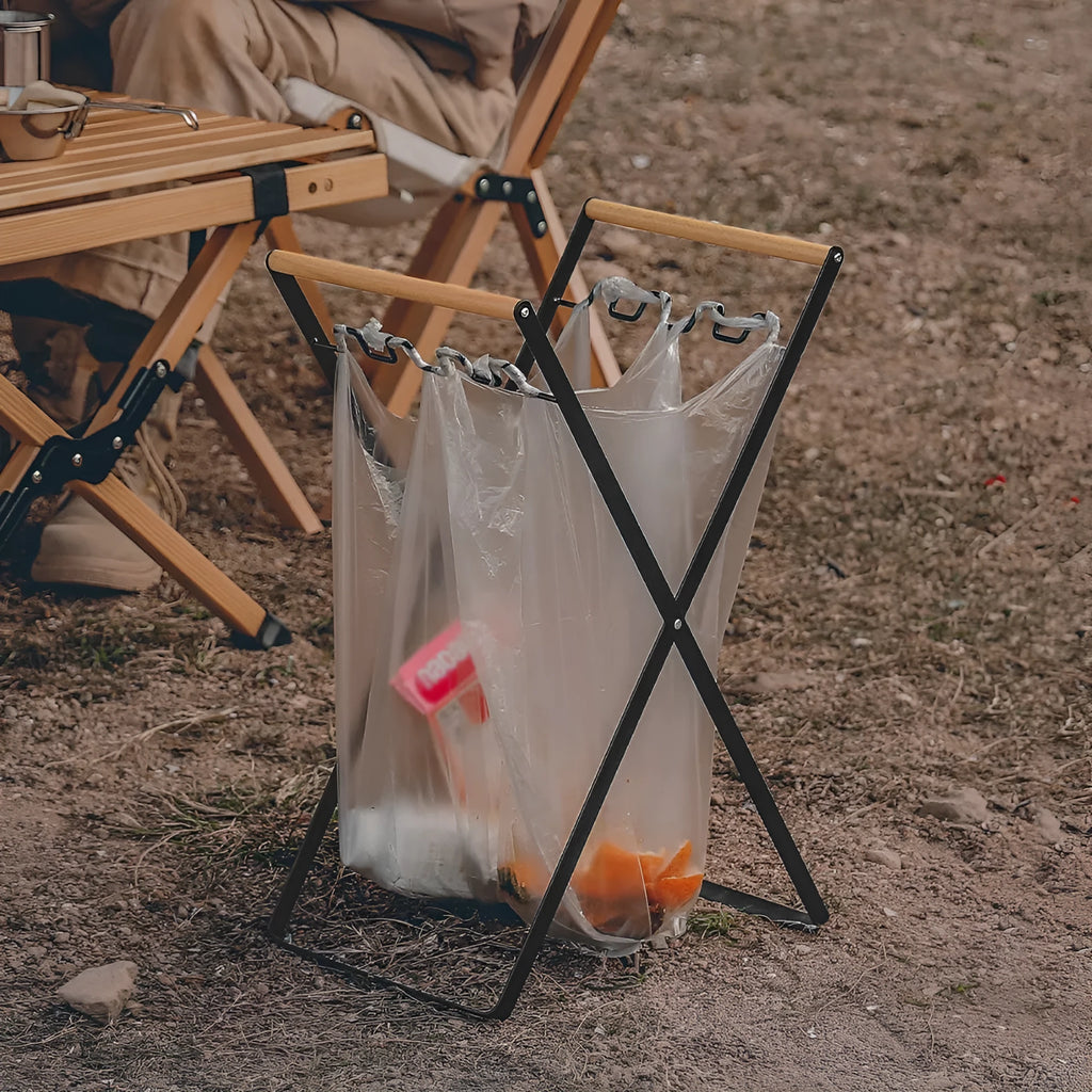 Black folding iron trash rack with wooden handle holding a bin bag outdoors.