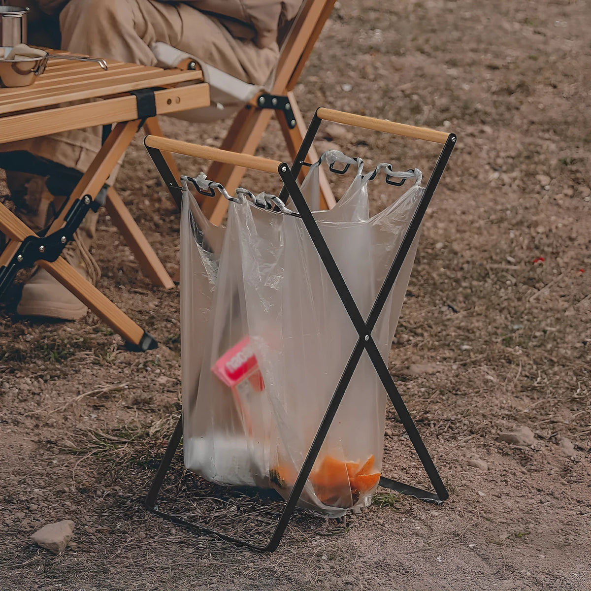Black folding iron trash rack with wooden handle holding a bin bag outdoors.